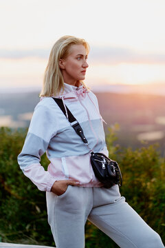 A thoughtful young blonde woman in casual sportswear and a crossbody bag gazes toward a warm sunset over rolling countryside, conveying calm, reflection and outdoor lifestyle during golden hour.