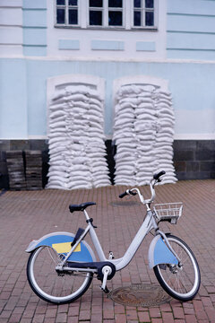A parked bicycle rests on a brick street before a pale blue building secured with stacked sandbags, suggesting urban flood protection, preparedness and quiet resilience on a rainy, overcast day.