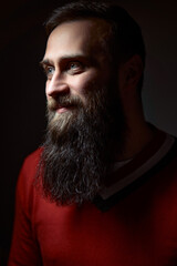 Profile portrait of a man with a full beard, lit dramatically, in a cozy environment. His red sweater accentuates his warm and confident demeanor, appealing to themes of masculinity and fashion. © Loginova