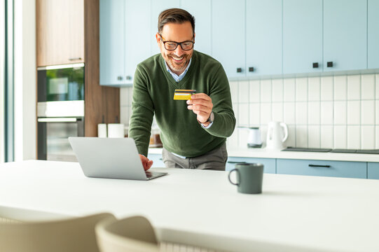 Smiling adult man standing in a modern kitchen setting, holding a credit card and using a laptop for secure online shopping and banking, making an electronic payment