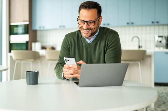 Professional man working remotely from a modern kitchen, using a mobile phone and laptop for business communication, accounting, and legal tasks