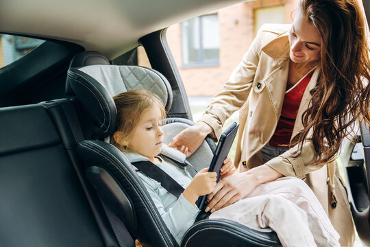 Mother securing child in car seat while daughter using tablet