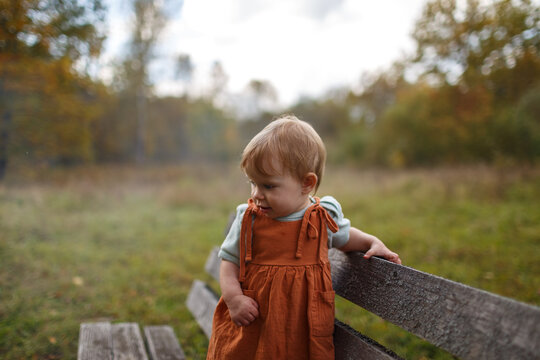 An infant under one year old stands next to a wooden bench in a park looking down