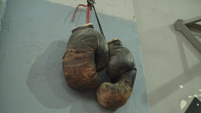 Pair of old and heavily worn boxing gloves hanging on a wall in a training gym, showing signs of intensive use and age.