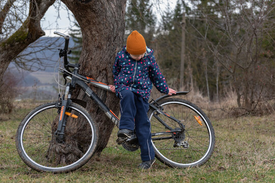 Young child in hat and jacket leans on an old tree trunk during a countryside bike ride, pausing to rest and enjoy a rural outdoor cycling adventure in cool weather