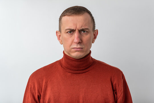 Man with short hair in a brown turtleneck stares at camera with a focused, frowning expression, conveying disbelief, skepticism, uncertainty and quiet frustration