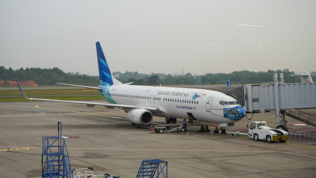 Garuda Indonesia Boeing 737-800 wearing mask livery parked at Hang Nadim Airport gate. Commercial aircraft serviced by ground crew and vehicles at the apron area.