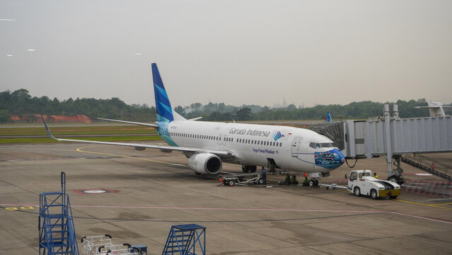 Garuda Indonesia Boeing 737-800 with special face mask livery parked at Hang Nadim International Airport tarmac connected to jet bridge during ground handling.