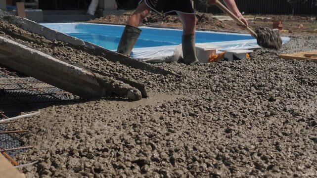 Construction workers pouring fresh concrete for pool deck area using mixer truck chute and shovels.