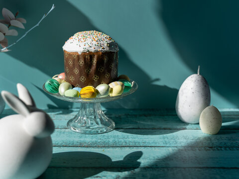 Traditional Paska Pasca Easter bread served on a glass cake stand with decorative Easter eggs and a white bunny ornament on a turquoise wooden background