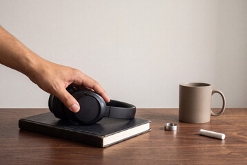 Fototapeta premium Hand lowering black over ear headphones onto a closed book beside a mug on a walnut desk