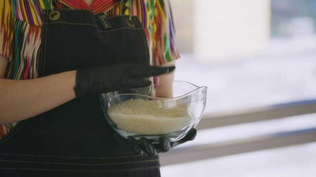 Young female chef in khan atlas holding glass bowl of rice for pilaf