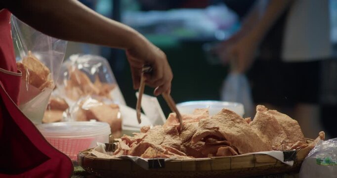 Street food vendor using tongs to pick and pack fried pork snacks into a plastic bag at a night market stall. Close-up of food preparation and takeaway service.