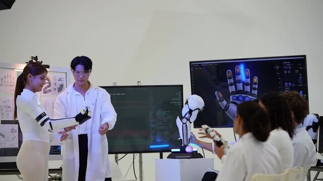 Woman with auburn ponytail extends a white robotic exoskeleton arm as an Asian engineer in goggles demonstrates it to a seated team, during a problem-solving session, a physical AI concept
