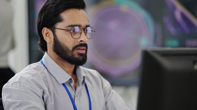 Bearded engineer with glasses and lanyard studies his monitor, then gestures as he talks to a colleague off screen under a glowing hologram display, physical AI
