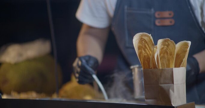 Street food vendor spreading crepe batter on a hot griddle with finished crepes displayed in a paper bag in the foreground at a night market stall.