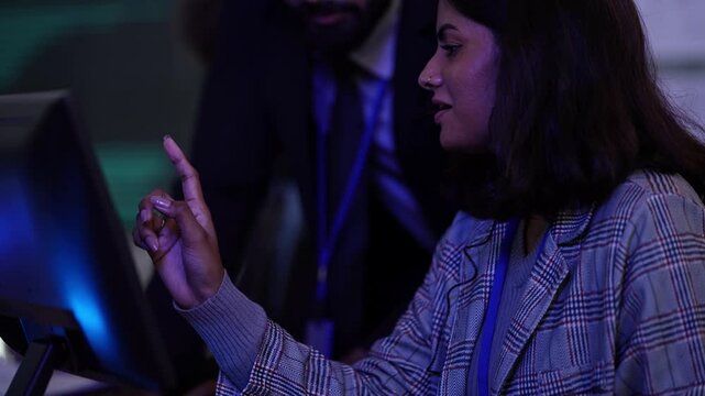 Curly-haired engineer with headset works as a bearded manager leans over an Indian female colleague in plaid blazer at a dark blue-lit workstation, in a candid moment, a physical AI concept