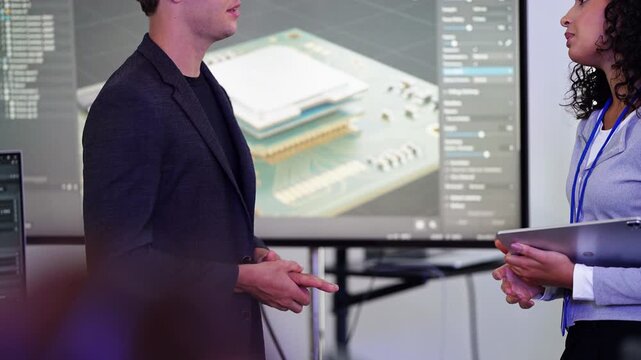 Young engineer in dark blazer rubs his hands and speaks as a curly-haired colleague holding a tablet looks on in front of a labeled layered chip design, real time
