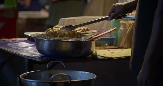 Close-up of a vendor deep frying seafood rolls at a Thai night market stall. Cooking process with hot oil, pan, and street food preparation in an outdoor setting.