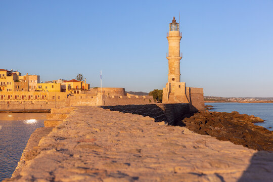 Historic Venetian lighthouse and stone harbor wall illuminated by warm sunrise light in Chania, Crete, Greece.