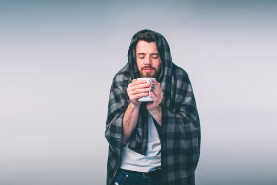 wrapped in the warm blanket or plaid, sick, young snotty man in glasses drinking warm healing tea