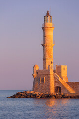 Fototapeta premium Historic Venetian lighthouse and stone harbor wall illuminated by warm sunrise light in Chania, Crete, Greece.