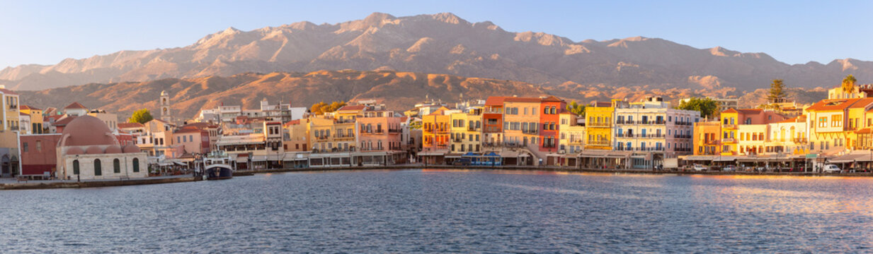 Colorful waterfront buildings and the historic harbor of Chania illuminated by warm sunrise light with the White Mountains in the background, Crete, Greece.