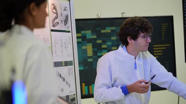 Seated curly-haired engineer with glasses points a pen at a bionic arm held by a colleague in front of a data heat map screen, during a collaborative session, an embodied AI research scene