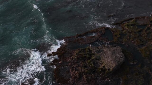 Dramatic aerial view of dark rocky coastline and foamy surf crashing against outcrops at Kelecung Beach, Tabanan, Bali, highlighting motion, raw ocean power and textured coastal patterns from above