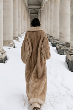 A woman in a long beige fur coat walks through a snow-covered classical colonnade, photographed from behind in winter.