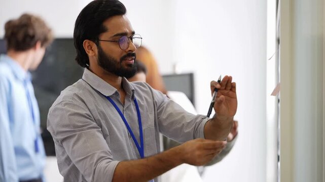 Bearded engineer gestures with a pen as an Indian female colleague in glasses leans in to check the tablet with him, coworkers passing behind, physical AI concept