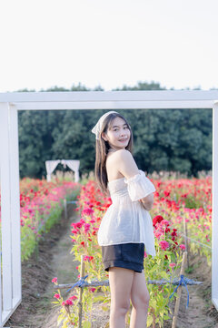 Beautiful young Asian woman standing inside a white wooden archway in a vibrant cockscomb flower garden, wearing a white top and shorts with a headscarf during sunny outdoor travel vacation in nature