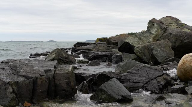 Rocky coastline with gentle ocean waves splashing against dark, jagged rocks along Englishman Bay at Roque Bluffs State Park in Washington County, Maine.