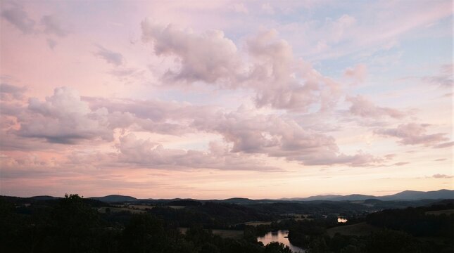 Soft pink and lavender sky fills the horizon. Clouds drift gently above rolling green hills. Calm river reflects the fading daylight. Distant trees frame the peaceful countryside