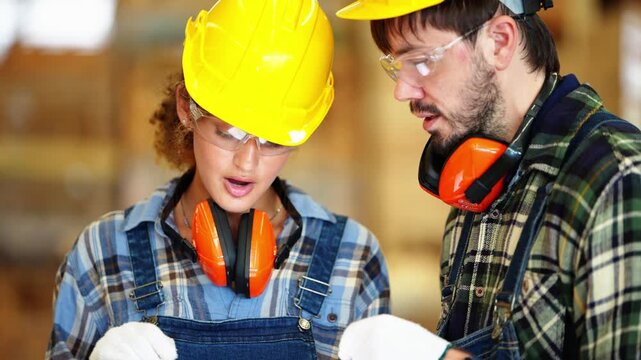Close portrait of a young female worker explaining a report on a tablet with a stylus as her bearded colleague leans in to read it, inside a plywood and timber factory