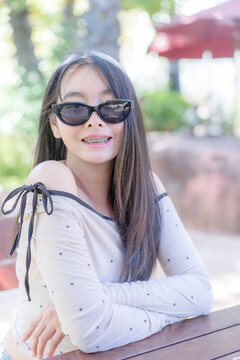 Close up portrait of a trendy young Asian woman wearing stylish black sunglasses and a polka dot top, smiling happily while resting her arms on a wooden table outdoors during a sunny leisure day