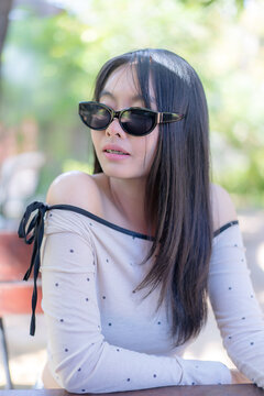 Portrait of a trendy young Asian woman wearing stylish black sunglasses and a polka dot top, looking away outdoors during a sunny leisure day in a natural background setting with soft lighting.