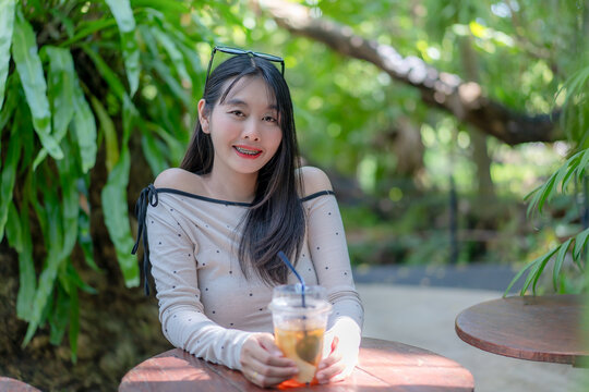 Portrait of a trendy young Asian woman holding an iced drink at a wooden table outdoors during a sunny leisure day, smiling with eyeglasses on her head in a lush green natural background setting