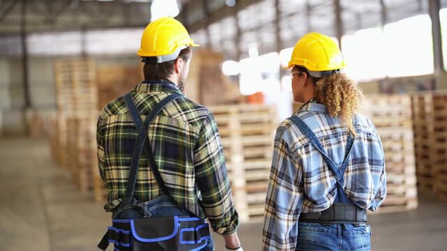 Rear view of a bearded man and a curly-haired woman in yellow hard hats walking deeper into a pallet warehouse between tall wooden stacks, in a timber factory, 4K