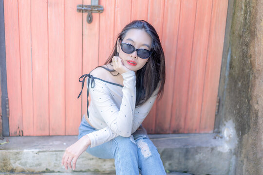 Portrait of a trendy young Asian woman wearing stylish black sunglasses and a polka dot top, resting her chin on her hand while sitting against a vibrant pinkish-orange wooden door background