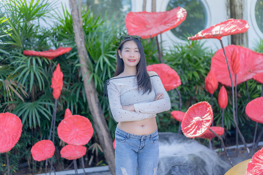 Portrait of a trendy young Asian woman crossing her arms and smiling amidst giant red lotus sculptures in a lush green garden during a sunny day, wearing a polka dot top and denim jeans.