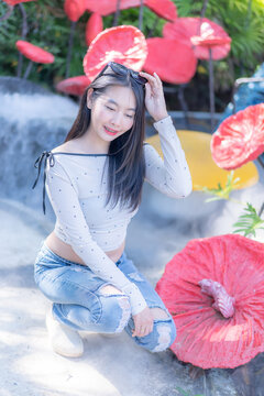 Portrait of a trendy young Asian woman squatting and adjusting her sunglasses amidst giant red lotus sculptures in a lush green garden during a sunny day, wearing a polka dot top and denim jeans