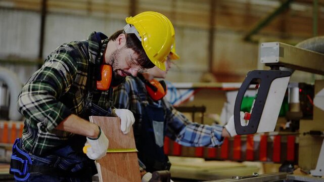 Female woodworker points to the saw blade while her bearded colleague lifts a plywood panel and stretches a tape measure across it inside a timber and plywood factory
