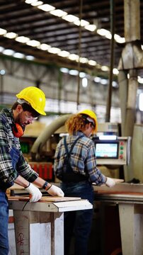 Vertical wide shot of a bearded carpenter cutting boards on a table saw while a female colleague programs a CNC touchscreen under long rows of lights, real time
