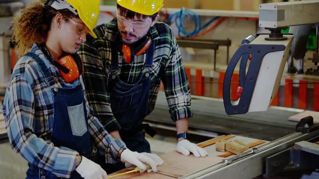Bearded supervisor points to the control arm of a panel saw as a curly-haired female trainee holding a tape measure leans in to listen, sustainable wood industry