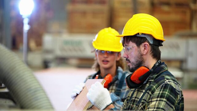 Close shot of a curly haired woman and a bearded man in yellow hard hats talking over a piece of plywood, then turning back to the machine, 4K video in a wood factory