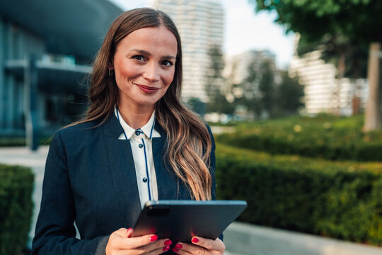 Businesswoman holding a tablet, looking at the camera and smiling, standing outdoors in an urban park