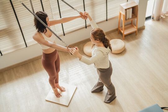 Pilates instructor and her client reach overhead together, both pulling resistance handles in perfect sync. This close-up moment captures shared focus, guided technique, and the connection between coa