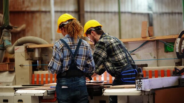 Rear view of a curly-haired woman and a bearded man in yellow hard hats leaning over a large panel saw to line up a board by weathered corrugated walls, real time