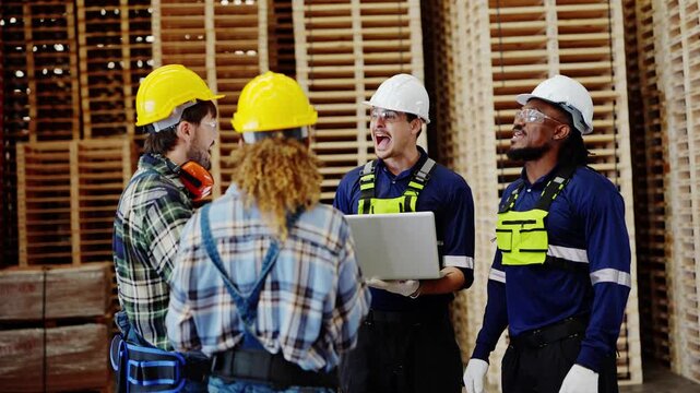 Team of four workers laughs together in a pallet warehouse as a Latino engineer holding a laptop smiles and a bearded coworker talks, inside a plywood and timber factory